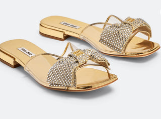 Planet Luxury Brands imzalı, kristal taş işlemeli fiyonk detaylı, altın rengi düz taban Miu Miu kadın sandalet.