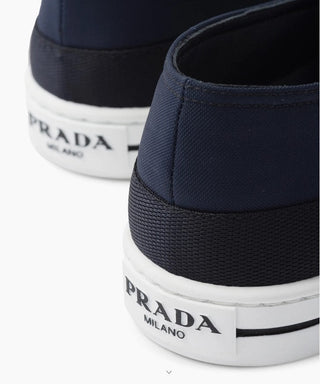 Planet Luxury Brands Prada lacivert Re-Nylon erkek bağcıksız ayakkabı, topuk kısmında beyaz kabartma logo detayı ile.