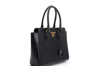 Planet Luxury Brands siyah Saffiano dana derisi Prada tote çanta, altın logo detaylı ve fermuarlı iç cepli tasarım.